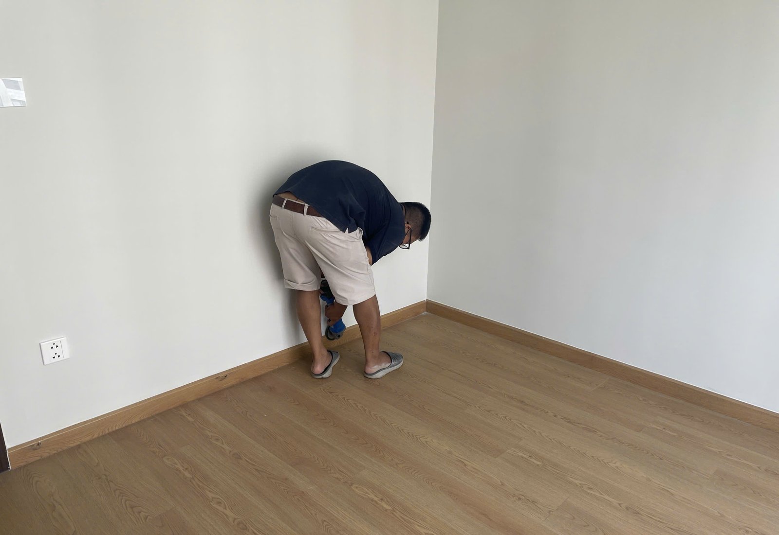 Người kiểm tra cúi sát góc tường và chân tường trong một phòng đã hoàn thiện, soát từng vị trí trước khi nhận bàn giao