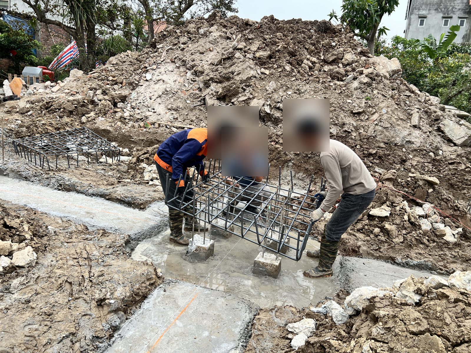 Ba người đang đưa lưới thép đài móng vào vị trí trên lớp bê tông lót; trên mặt lót giằng ở tiền cảnh có một vạch màu cam và một sợi dây căng