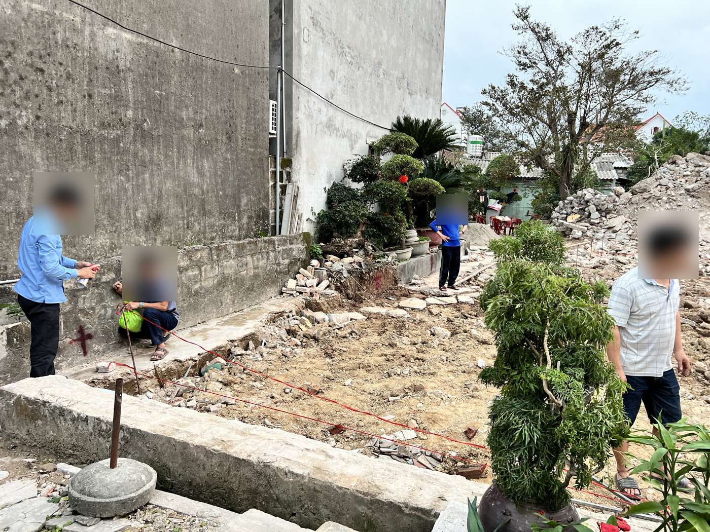 Cọc mốc thép cắm trong khối bê tông tròn đặt ở góc thửa đất, dây trục đỏ chạy dọc chân tường nhà bên cạnh, trên tường có dấu sơn đỏ đánh dấu