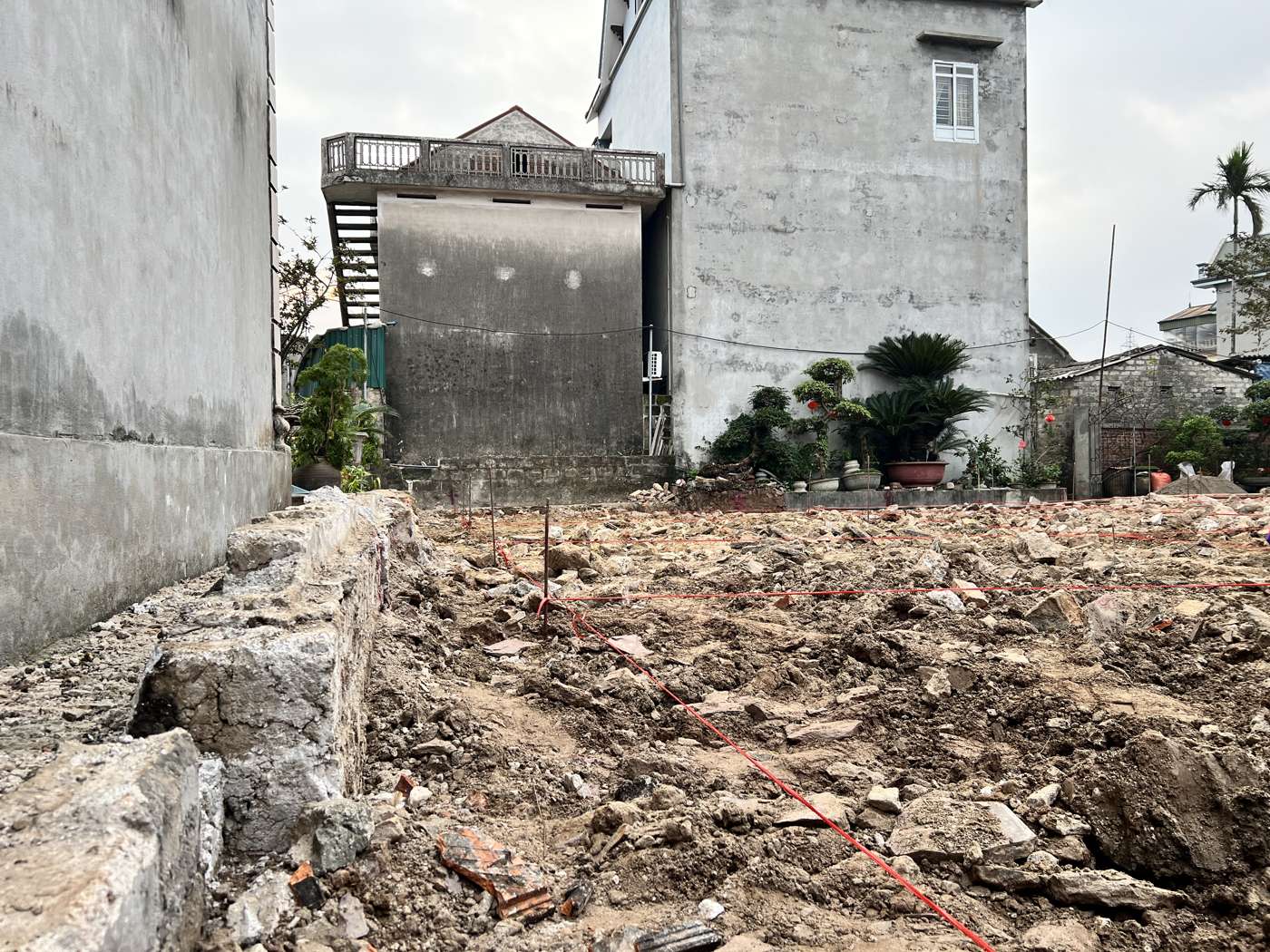 Dây trục màu đỏ căng trên cọc thép chạy dọc phía tường nhà bên cạnh, sát một khối đá xây cũ ở chân tường, phía sau là các nhà liền kề