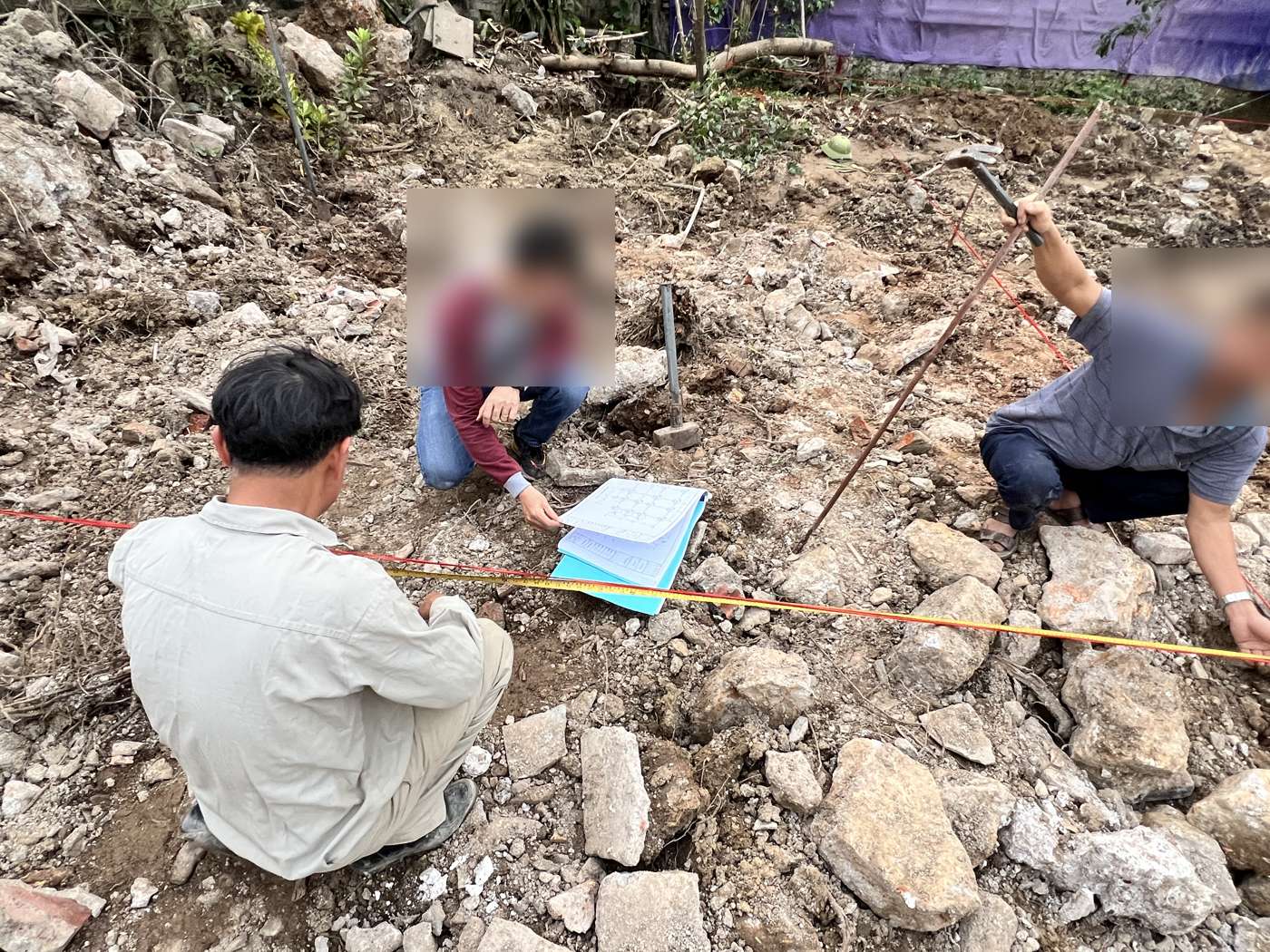 Ba người ngồi trên mặt bằng đất đá vừa dọn, một người cầm tập bản vẽ, hai người kéo thước dây vàng và dây căng đỏ để đo kiểm vị trí trục