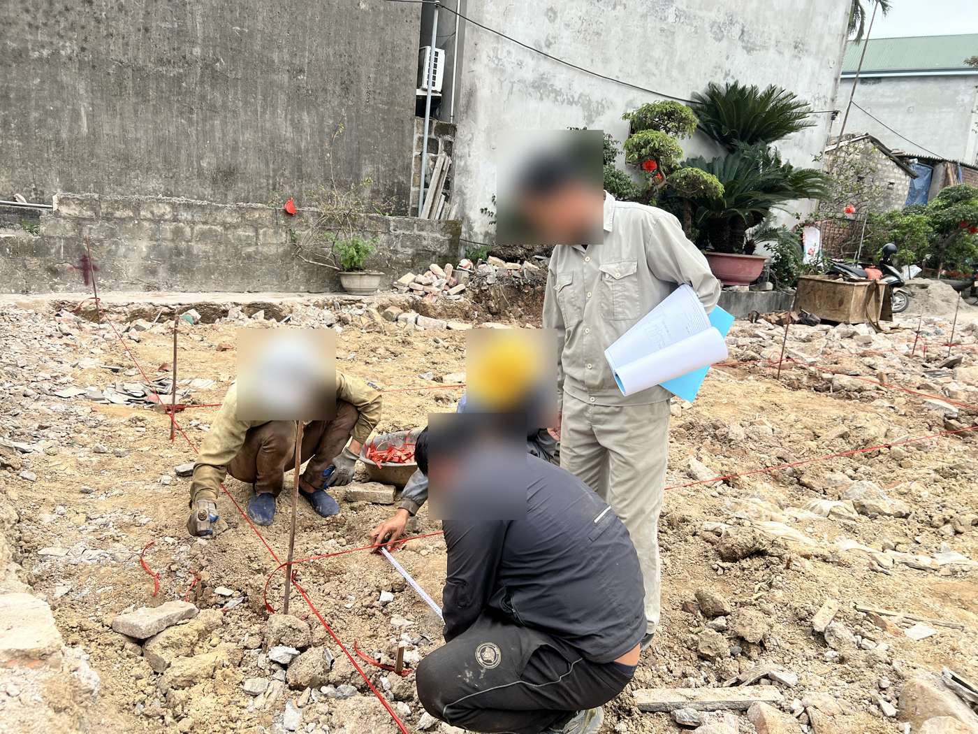 Nhóm người ngồi quanh giao điểm hai dây trục đỏ, một người đặt thước đo, người đứng bên cạnh cầm tập bản vẽ