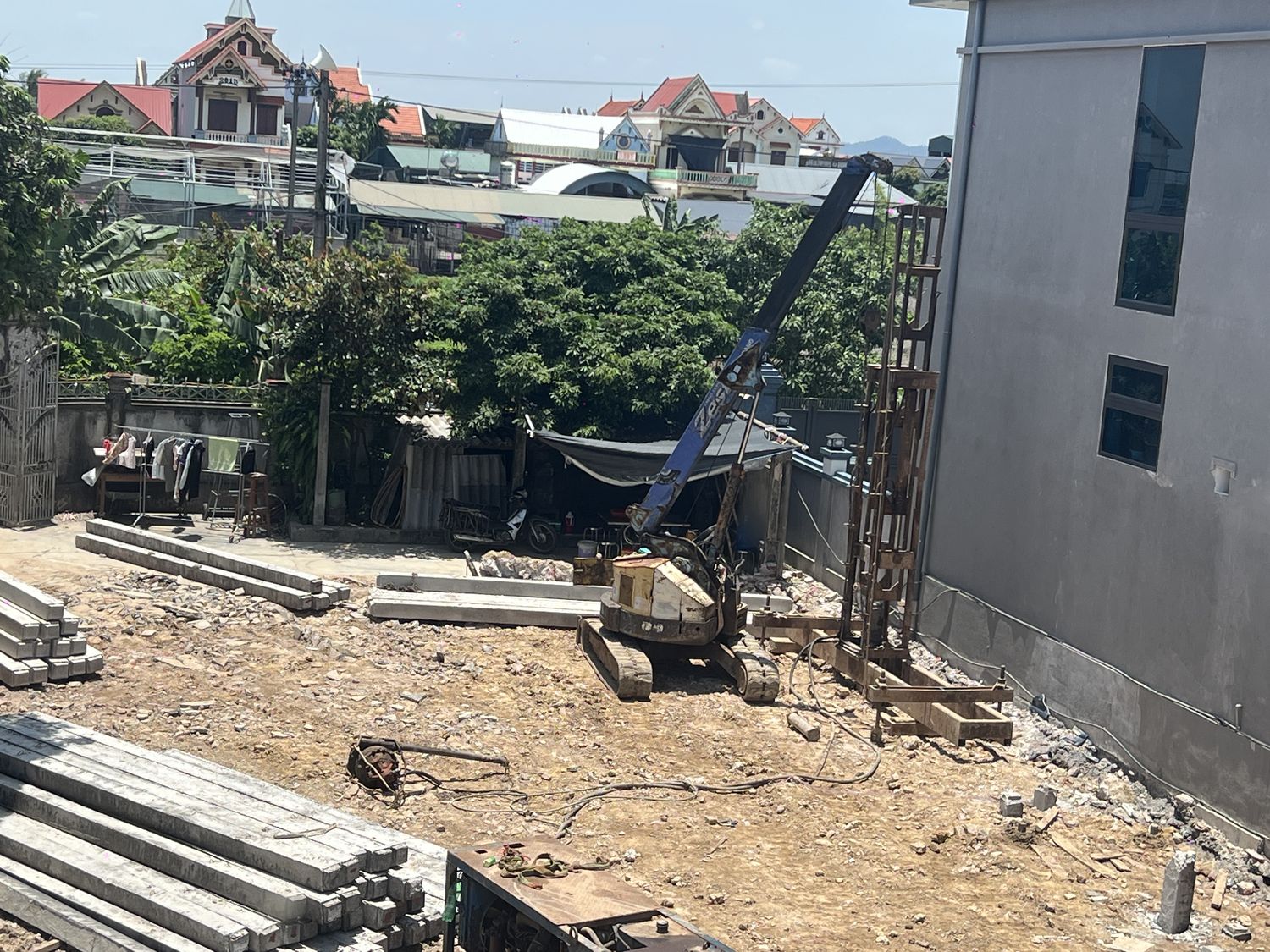 Công trường ép cọc nhà dân trong khu dân cư, cọc bê tông vuông tập kết thành từng chồng ở tiền cảnh, giá ép và cẩu bánh xích đứng sát tường nhà bên