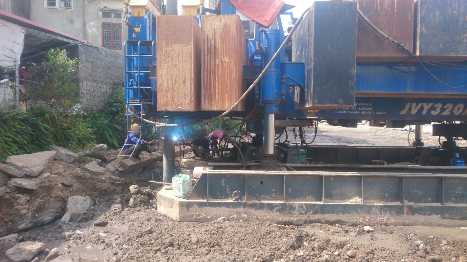 Thợ đang hàn nối hai đoạn cọc ngay tại vị trí ép, tia lửa hàn sáng rõ dưới thân máy ép cọc, tường nhà dân ngay phía sau