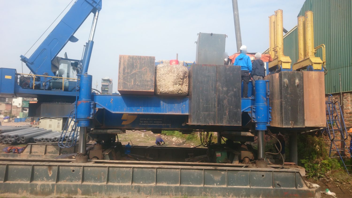 Máy ép cọc thủy lực tự hành với hệ đối trọng gồm nhiều khối thép và bê tông xếp trên khung máy, cẩu bánh xích đứng cạnh