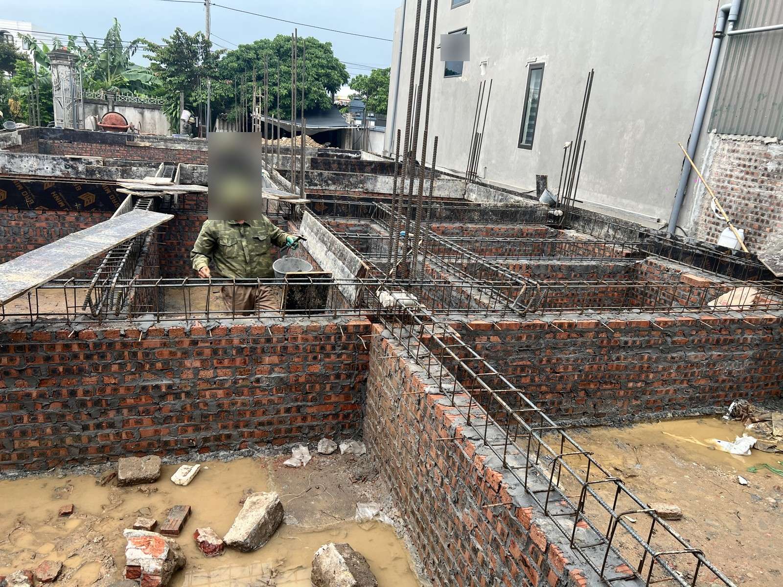 Đáy hố quanh móng xây gạch nhà dân còn đọng nước bùn, gạch vỡ và đá rơi vãi, thợ đang làm việc phía trên giằng móng