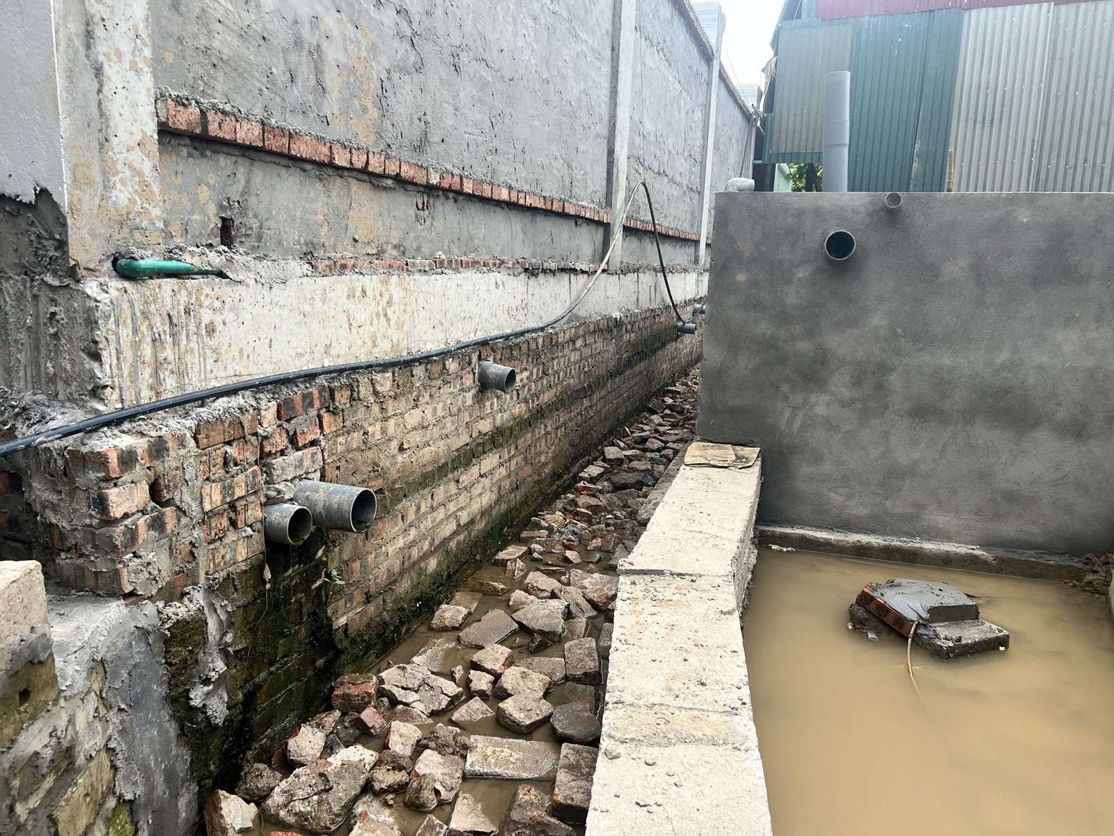 Chân tường nhà bên cạnh lộ ra khi làm móng, có các đầu ống thoát nước nhô ra ở chân tường, hố bên cạnh đang đọng nước đục