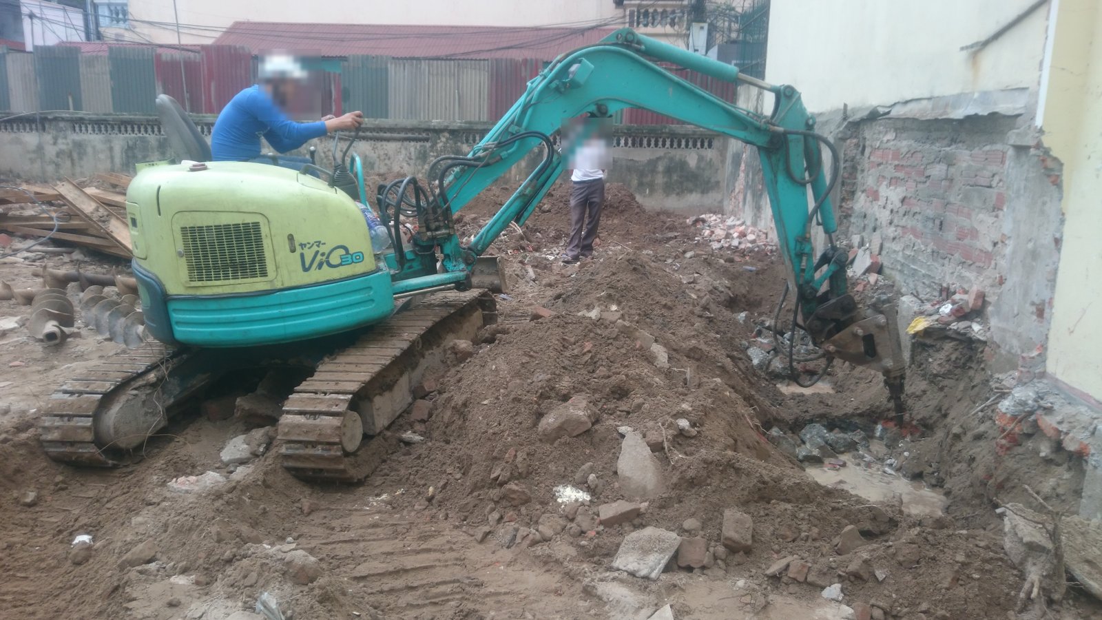 Máy đào mini đang phá đất sát chân tường nhà bên, phần gạch cũ ở chân tường nhà bên đã lộ ra