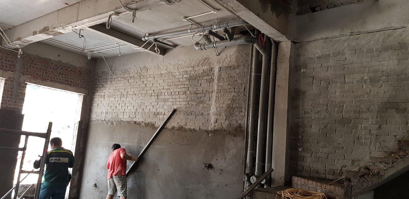 Thợ đang dùng thước nhôm dài cán phẳng lớp vữa trát trên tường gạch, phần tường phía trên chưa trát