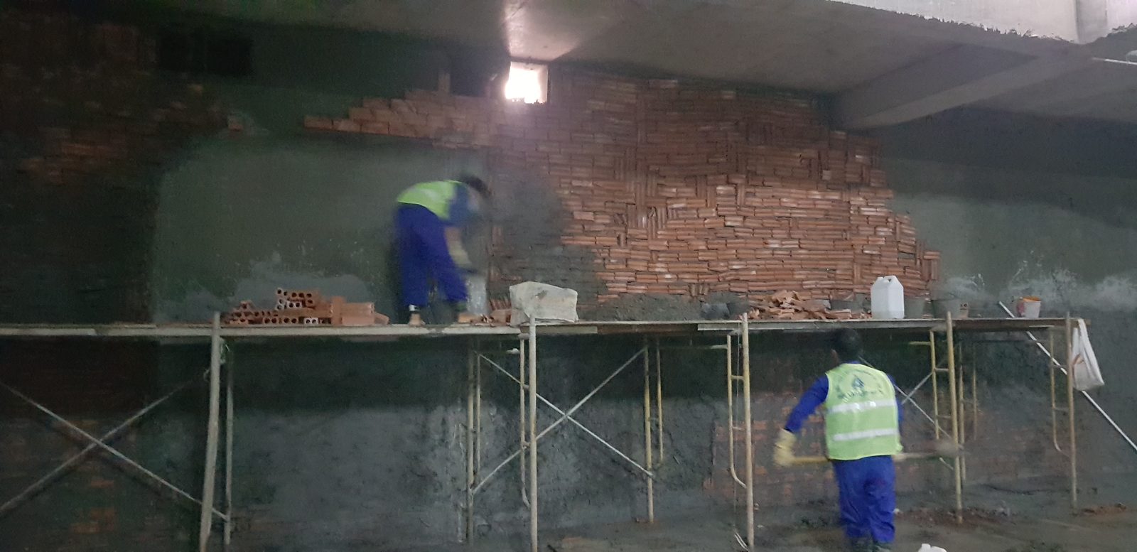 Hai thợ xây đang xây phần tường gạch phía trên giáp đáy dầm, đứng trên hệ giàn giáo thép