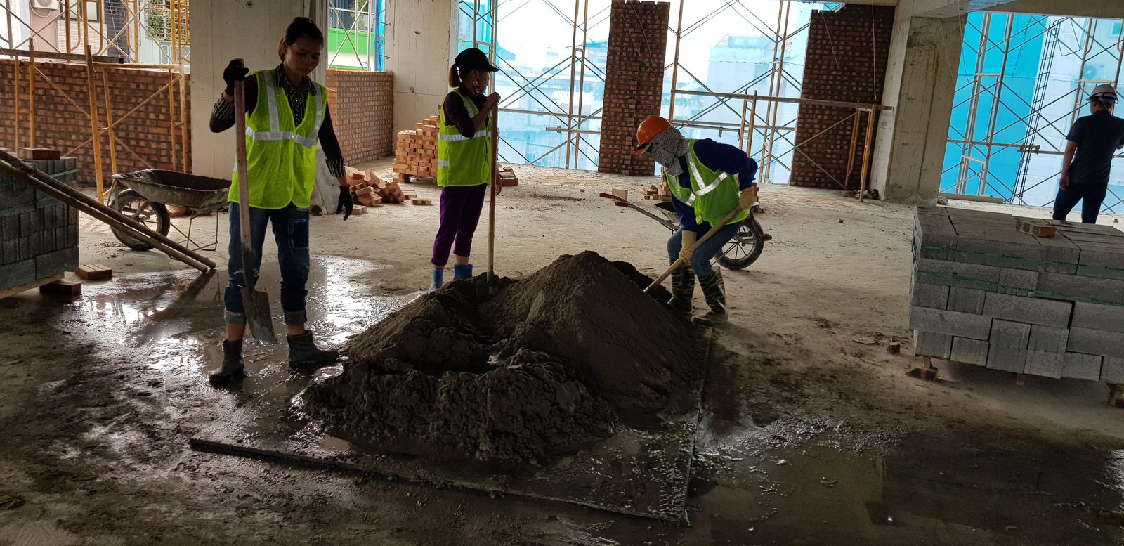 Ba công nhân đang trộn vữa xây bằng xẻng trên mặt sàn bê tông công trường, phía sau là gạch tập kết