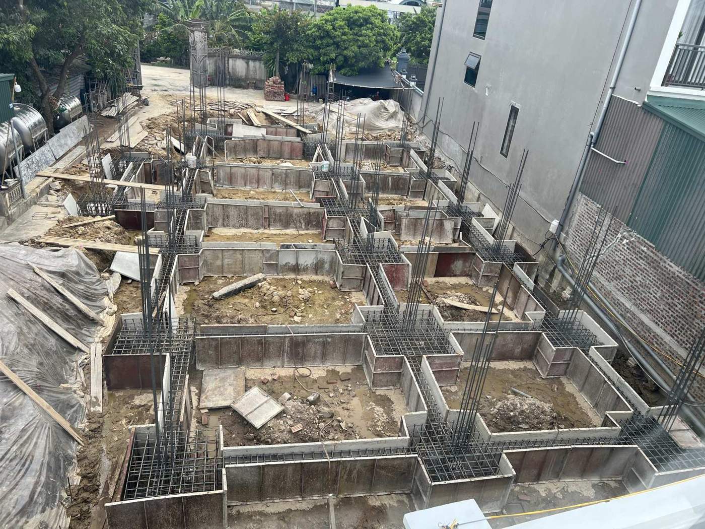 Toàn cảnh hệ cốp pha đài và giằng móng của một công trình nhà dân, thép cột chờ dựng đứng, lòng khuôn còn đất và nước đọng chưa vệ sinh