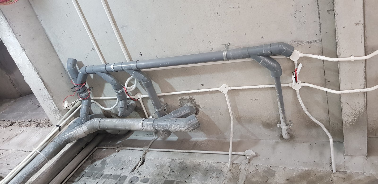 Ống thoát nước bằng nhựa xám và tuyến ống điện màu trắng cùng chạy trên trần và tường, chưa bị che kín