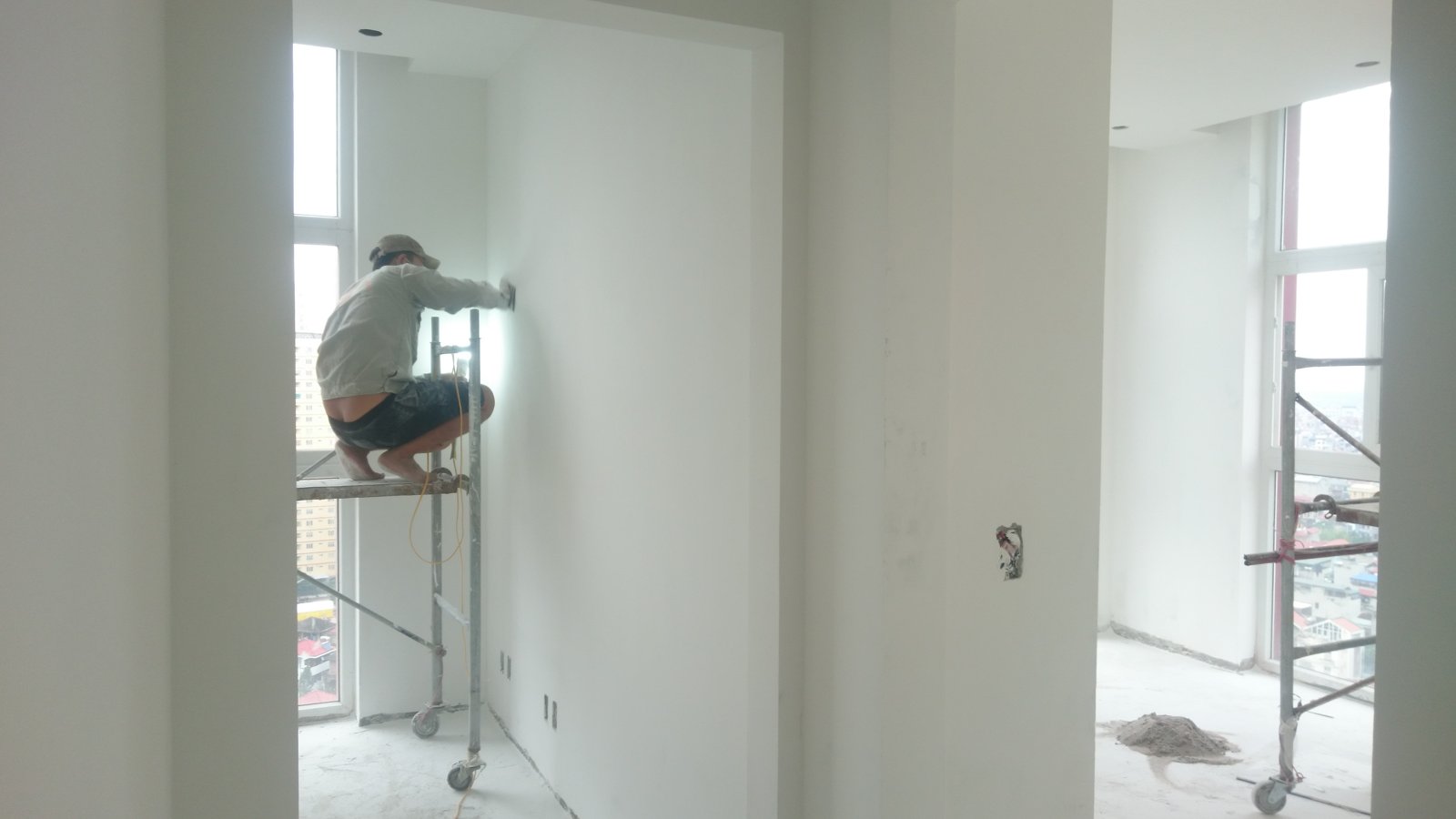 Thợ ngồi trên giàn giáo di động xử lý bề mặt tường đã bả ở góc cạnh cửa sổ