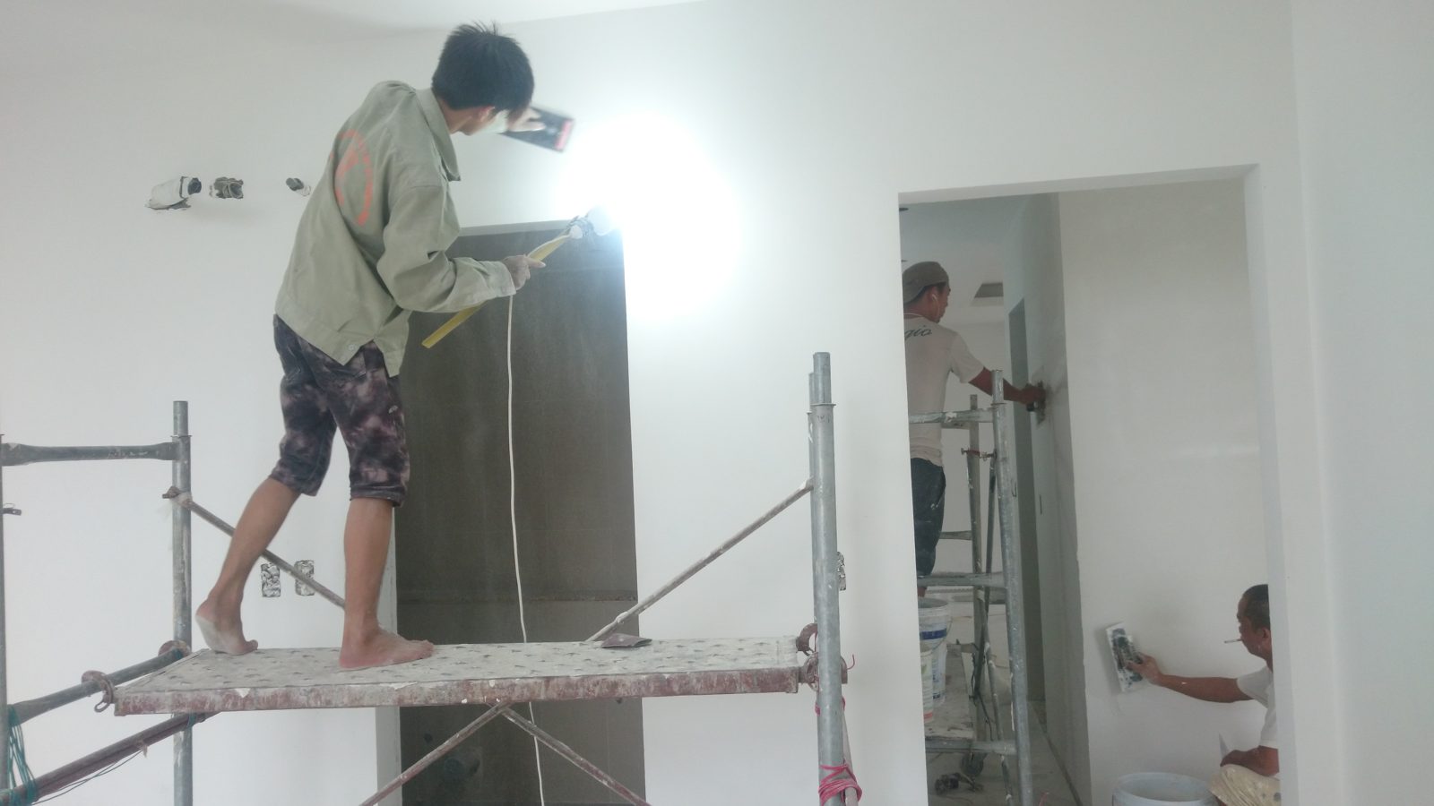 Thợ hoàn thiện soi đèn rọi men theo mặt tường để phát hiện gợn sóng và vết hằn trước khi sơn
