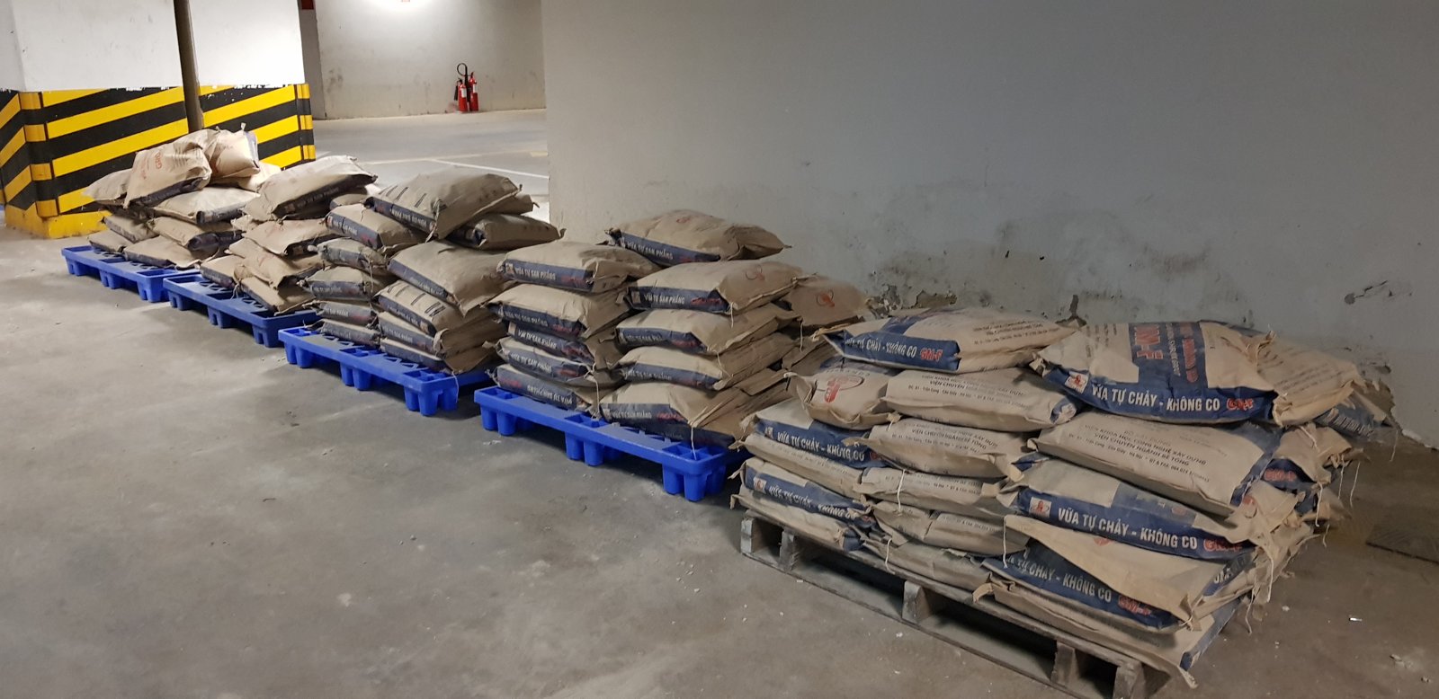 Các bao vữa khô xếp thành hàng trên pallet nhựa và pallet gỗ, kê cao khỏi mặt nền bê tông trong khu vực có mái che